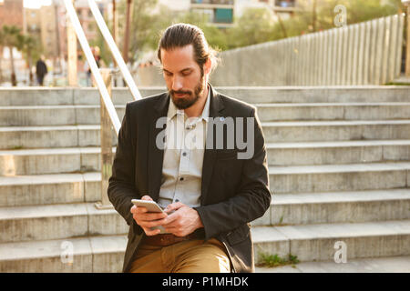 Photo of serious businesslike man 30s in formal suit using mobile phone while walking through town and standing on stairs in urban area Stock Photo