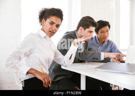 Confused young african woman sitting at the table with colleagues and working Stock Photo