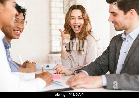 Smiling young businesswoman sitting at the table with her colleagues during meeting and showing thumbs up Stock Photo