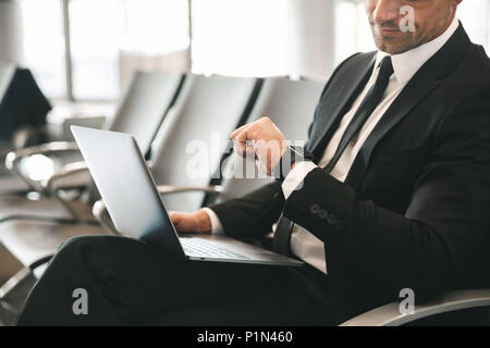 Cropped image of businessman dressed in suit using laptop computer and checking time while sitting at the airport lobby Stock Photo