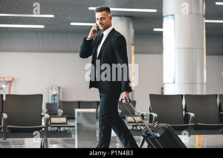 Smiling mature businessman talking on mobile phone while walking at the airport lobby with a suitcase Stock Photo