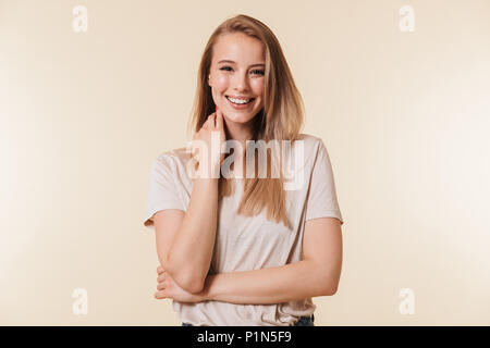 Portrait of charming blonde woman 20s wearing casual t-shirt smiling at camera while touching neck isolated over beige background in studio Stock Photo
