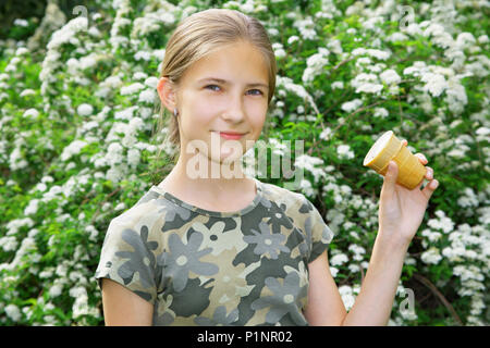 A pretty 13 year old girl with ice cream in her hand smiles at the camera. In the background, bushes of bird cherry blooming Stock Photo