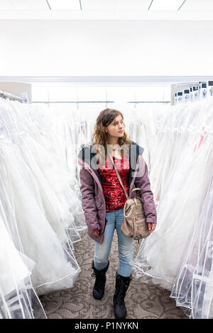 Young woman shopping for wedding dress gowns in boutique discount store aisle, purse, many white garments hanging on rack hangers row Stock Photo