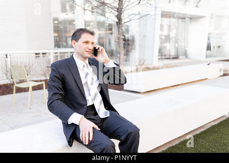 Funny, angry, irritated, confused businessman sitting on bench making face talking holding smartphone phone mobile cellphone in suit and tie on interv Stock Photo