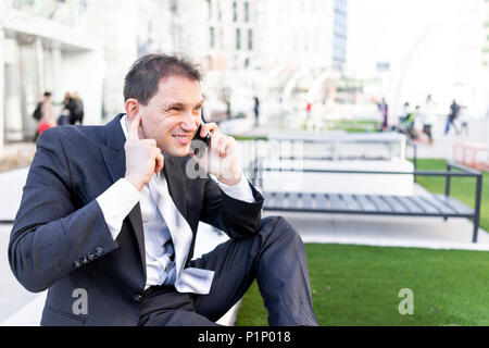 Funny businessman sitting on bench making face hearing impaired talking holding smartphone phone mobile cellphone in suit and tie on interview break i Stock Photo