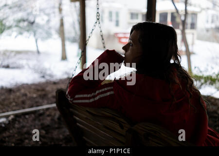 Closeup of young woman sitting on swing under wooden deck of house on backyard with snow during blizzard white storm, snowflakes falling in Virginia Stock Photo
