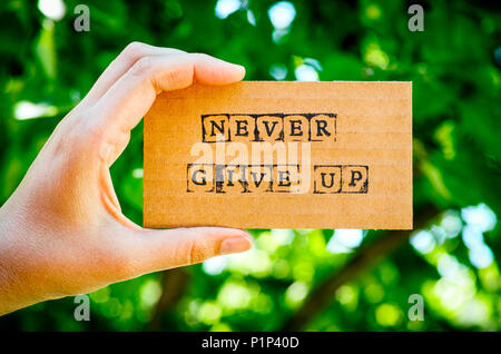 Woman hand holding cardboard card with words Never Give Up made by black alphabet stamps against green nature background. Stock Photo
