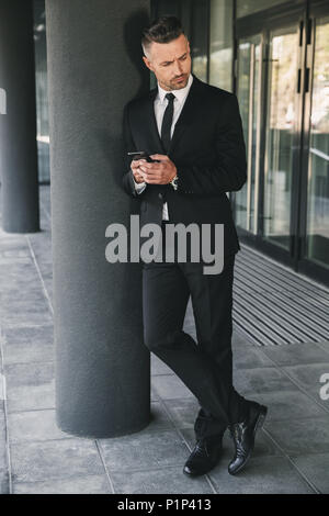 Portrait of a concentrated businessman dressed in suit standing outside a glass building and using mobile phone Stock Photo
