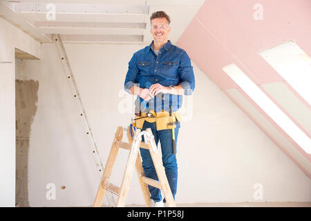Portrait of smiling construction man wearing protective eye googles while standing at the ladder and holding a drill in his hand. Stock Photo