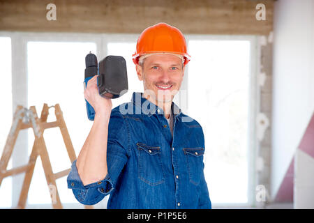 Smiling handyman wearing safety helmet and holding drill in his hand while standing at construction site. Stock Photo