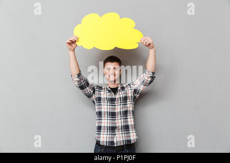 Portrait of a smiling young man in plaid shirt showing blank screen speech bubble over gray background Stock Photo