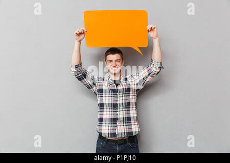 Portrait of a happy young man in plaid shirt showing blank screen speech bubble over gray background Stock Photo