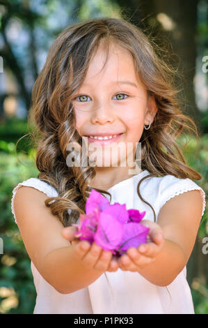 Portrait of a Caucasian five years old girl smiling and offering purple flowers in a park as a gift Stock Photo