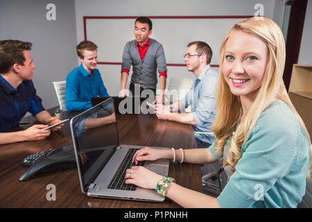 Young millennial business woman posing for the camera while a co-worker is speaking to a group of her piers in a conference room Stock Photo