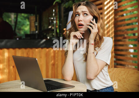 Portrait of a surprised young girl talking on mobile phone while sitting with laptop computer at a cafe outdoors Stock Photo