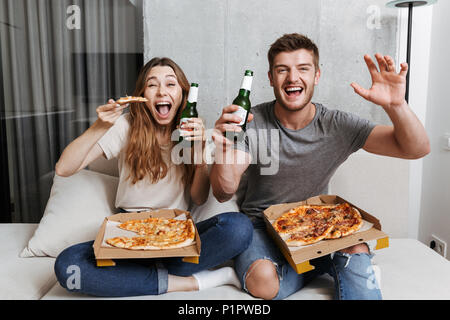 Excited young couple eating pizza and drinking beer while sitting together on couch at home and watching TV Stock Photo