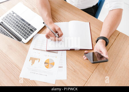 Cropped image from above of caucasian businessman sitting at table and writing down data or report at notebook from his mobile phone while working in  Stock Photo