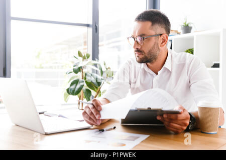 Portrait of smart businesslike man 30s in white shirt verifying information from paper documents using laptop during work in office Stock Photo