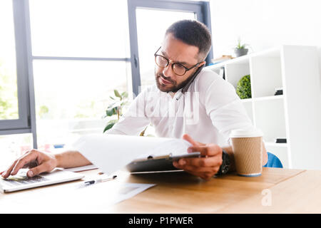 Portrait of smart businesslike man 30s in white shirt verifying information from paper documents using laptop and talking on cell phone during work in Stock Photo