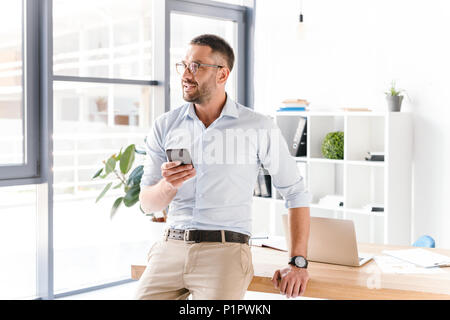 Horizontal photo of stylish man wearing glasses sitting on table in office and looking through big window while using mobile phone Stock Photo