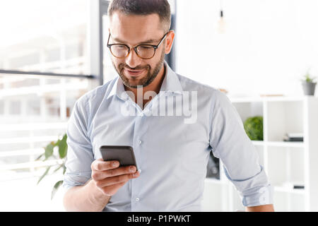 Image closeup of handsome employer man in formal wear standing in office near big window and using smartphone for work Stock Photo