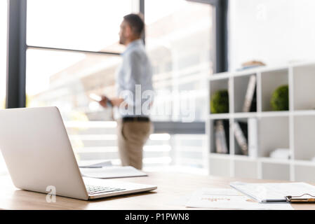 Photo of workplace with open white laptop lying on table while defocused business man standing and looking through big window on background Stock Photo