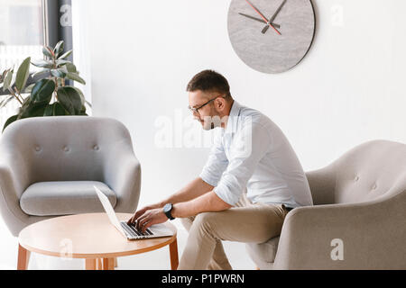Horizontal photo of business man wearing stylish formal clothing sitting in armchair, and working on laptop in office interior Stock Photo
