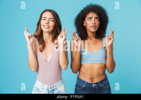 Two worried young girls dressed in summer clothes holding fingers crossed for good luck isolated over blue background Stock Photo