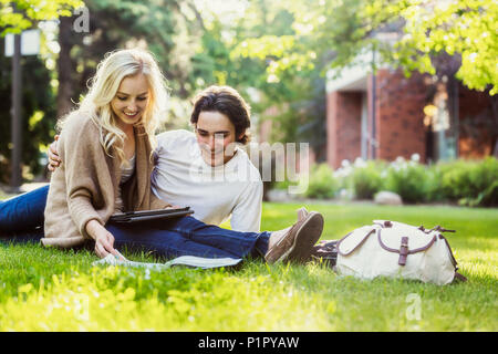 A young couple studying outdoors on the grass on the university campus and looking through a textbook while using a tablet; Edmonton, Alberta, Canada Stock Photo