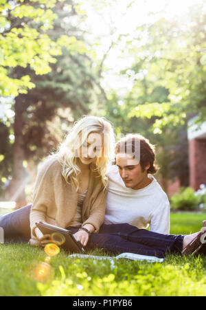 A young couple studying outdoors on the grass on the university campus and looking through a textbook while using a tablet; Edmonton, Alberta, Canada Stock Photo