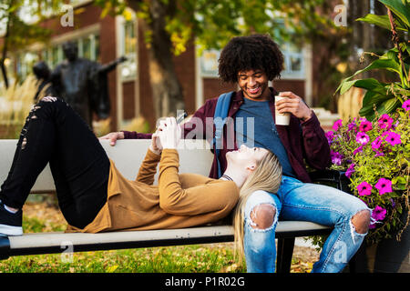Young dating couple who are university students relaxing on a bench on campus and the woman is checking her smart phone while laying her head on he... Stock Photo