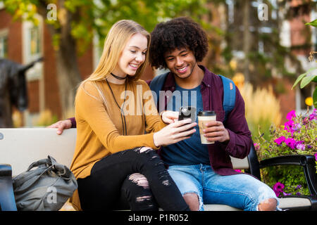 Young dating couple who are university students sitting on a bench on campus and looking at social media on a smart phone; Edmonton, Alberta, Canada Stock Photo