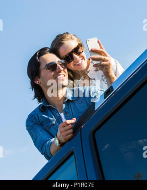 A young couple stand up through the sunroof of their vehicle and take a self-portrait with their cell phone; Edmonton, Alberta, Canada Stock Photo