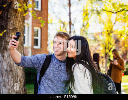 A couple who are university students posing for a self-portrait with a smart phone with a small group of friends in the background Stock Photo