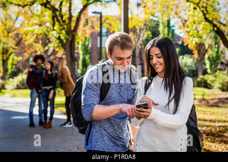 A male and female university student stand on a path looking at a smart phone together with a group of fellow students in the background Stock Photo