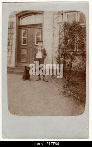 HAMBURG, GERMANY (DEUTSCHES REICH) - CIRCA 1920s: Vintage photo shows boy poses in front of house. Boy wears a sailor costume and stands next to dog. Retro black & white photography.pet Stock Photo