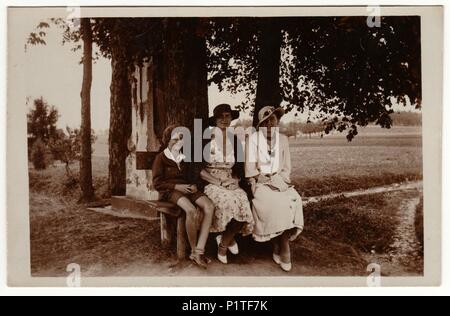 THE CZECHOSLOVAK REPUBLIC - CIRCA 1930s: Vintage photo shows women wear  elegant dresses and women's parisisol big brim hat. The women and boy pose outdoors and sit on the bench. Retro black & white photography. Stock Photo