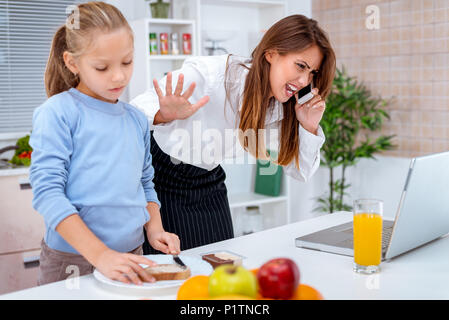 A little girl prepares a breakfast while her overworked mother phoning, and checking something on laptop, before she goes in the office. Mother is get Stock Photo