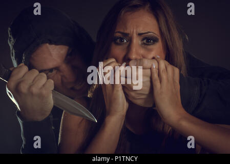 Young woman is abused victim by violence man who threatens with a knife and puts his hands on her mouth not to scream. Stock Photo