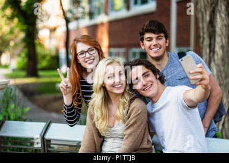 A group of four friends sitting on a bench taking a self-portrait with a smart phone on a university campus; Edmonton, Alberta, Canada Stock Photo