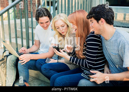 Four students sitting in a row on a step using their technology on the university campus and laughing together; Edmonton, Alberta, Canada Stock Photo