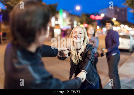 A young couple wearing black leather jackets being playful on a city sidewalk at night with friends watching in the background Stock Photo
