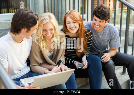 Four students sitting in a row on a step using their technology on the university campus and laughing together; Edmonton, Alberta, Canada Stock Photo
