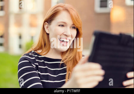 A young woman with red hair smiles big while looking at a tablet; Edmonton, Alberta, Canada Stock Photo