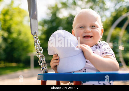 Baby boy playing in playground area. Portrait of smiling toddler looking at camera with happy face, having fun. Swaying on sunny summer day outdoors. Stock Photo