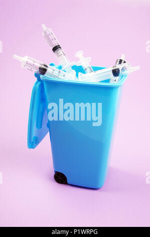 clear plastic medical syringes inside a blue rubbish bin with open lid on a pink tint background Stock Photo