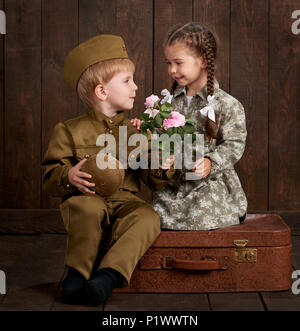 children boy are dressed as soldier in retro military uniforms and girl in pink dress sitting on old suitcase, dark wood background, retro style Stock Photo