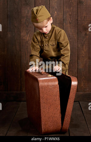 children boy are dressed as soldier in retro military uniforms with old suitcase, dark wood background, retro style Stock Photo