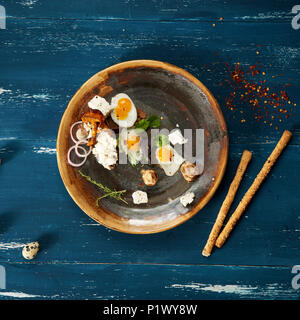 Bruschetta with cream cheese, fried eggs, mushrooms and salad leaves lying on plate standing on blue aged wooden surface with quail eggs and bread sti Stock Photo